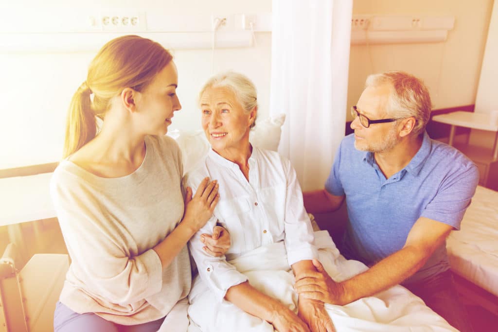 medicine, support, family health care and people concept - happy senior man and young woman visiting and cheering her grandmother lying in bed at hospital ward