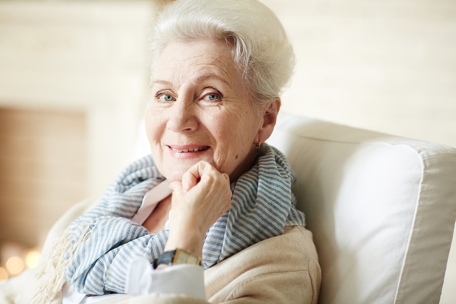 Portrait of beautiful old lady in beige cardigan and striped scarf sitting in armchair, looking at camera and smiling cheerfully